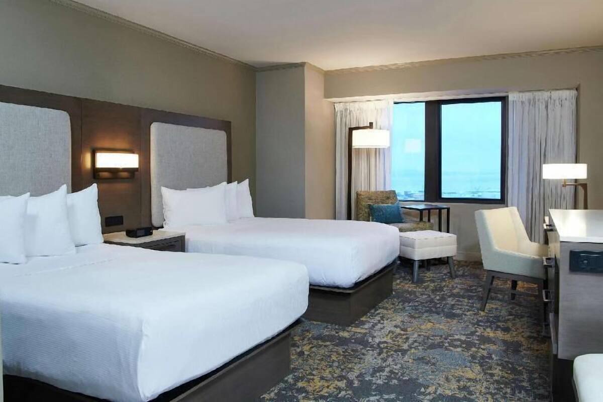 A hotel room with two beds, a desk, a chair, a small table, and a view of the ocean.