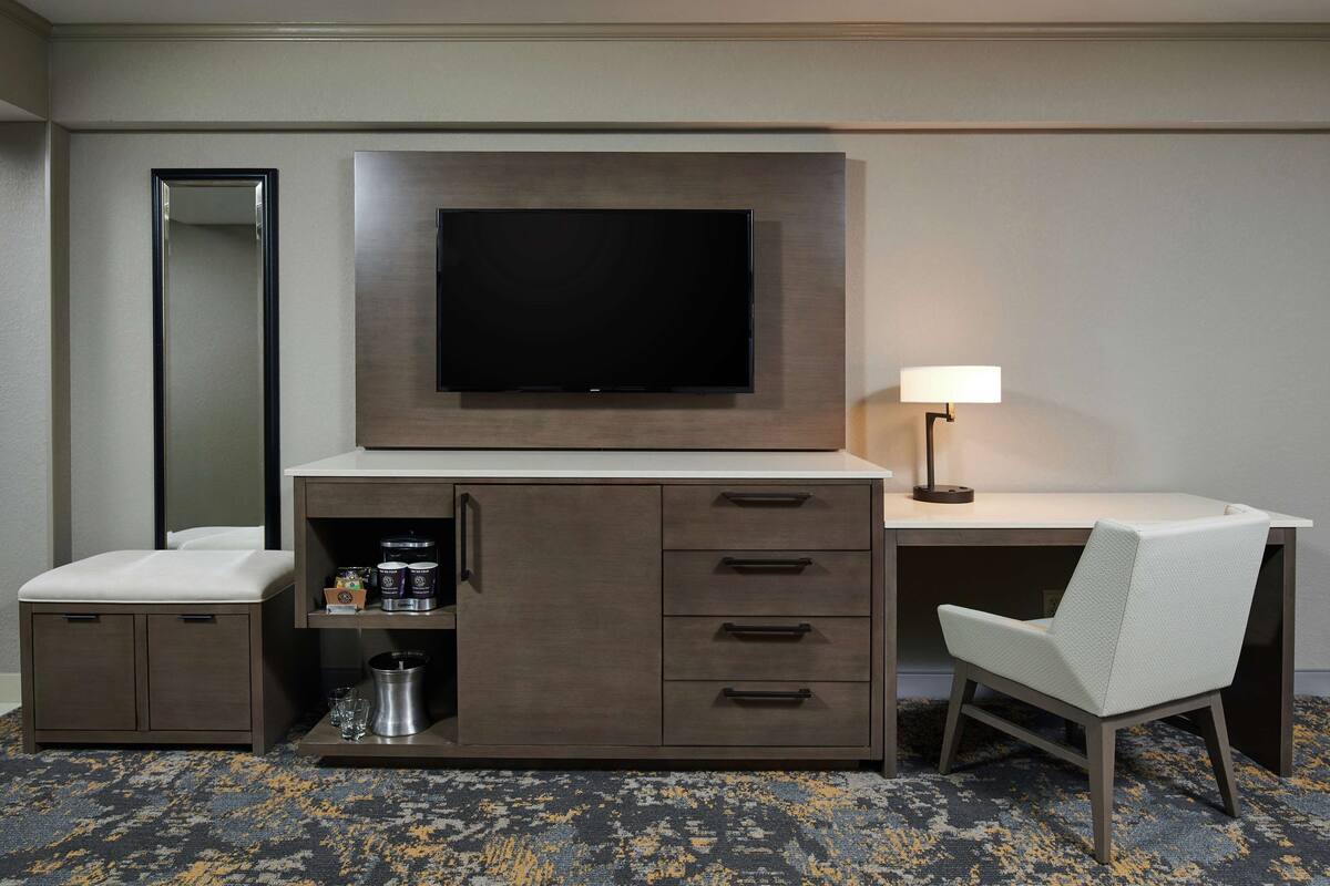 A hotel room with a flat-screen TV mounted on a cabinet, a desk with a lamp, a chair, and a small kitchenette area.