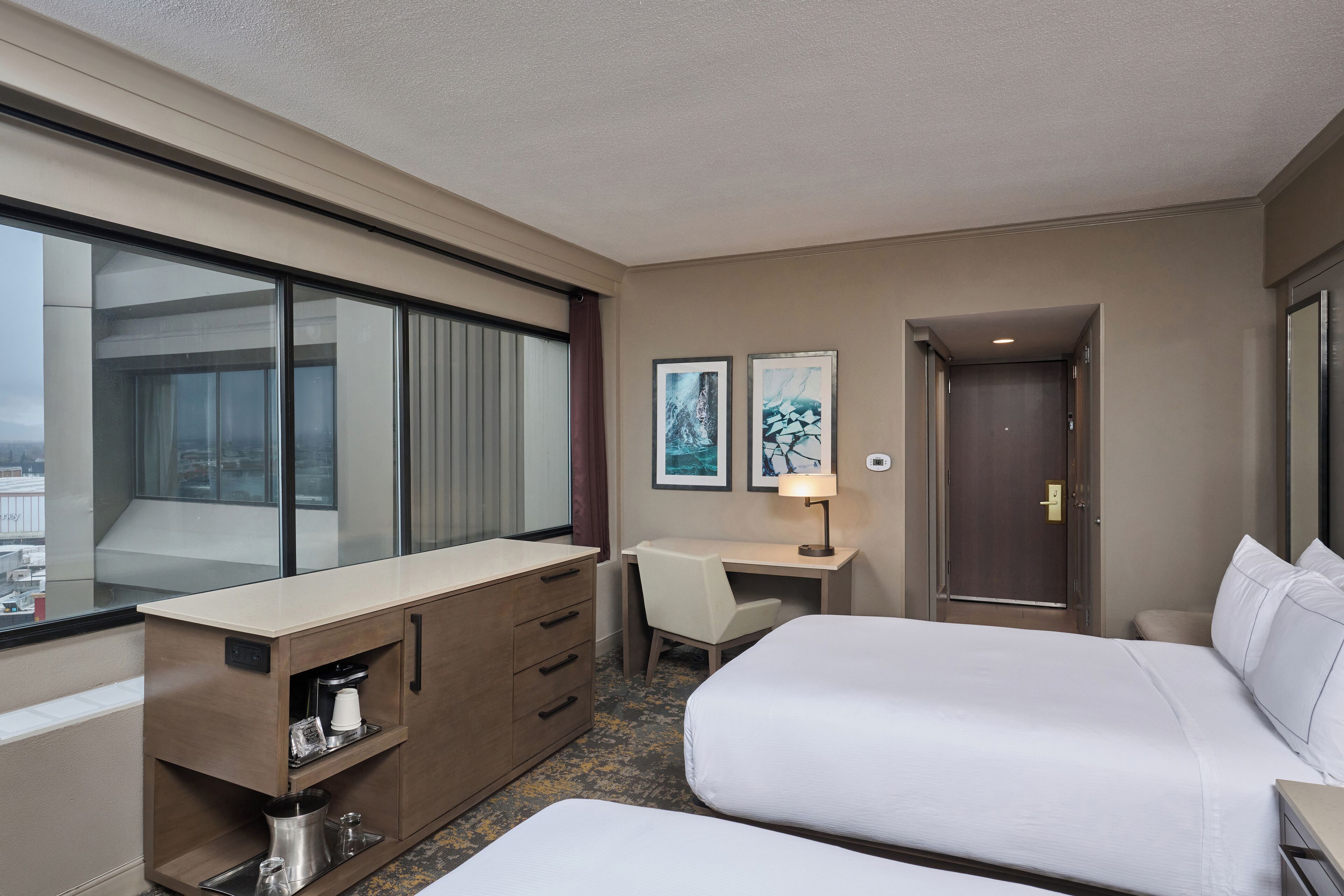 A hotel room with two beds, a desk, a chair, a minibar, and a large window.