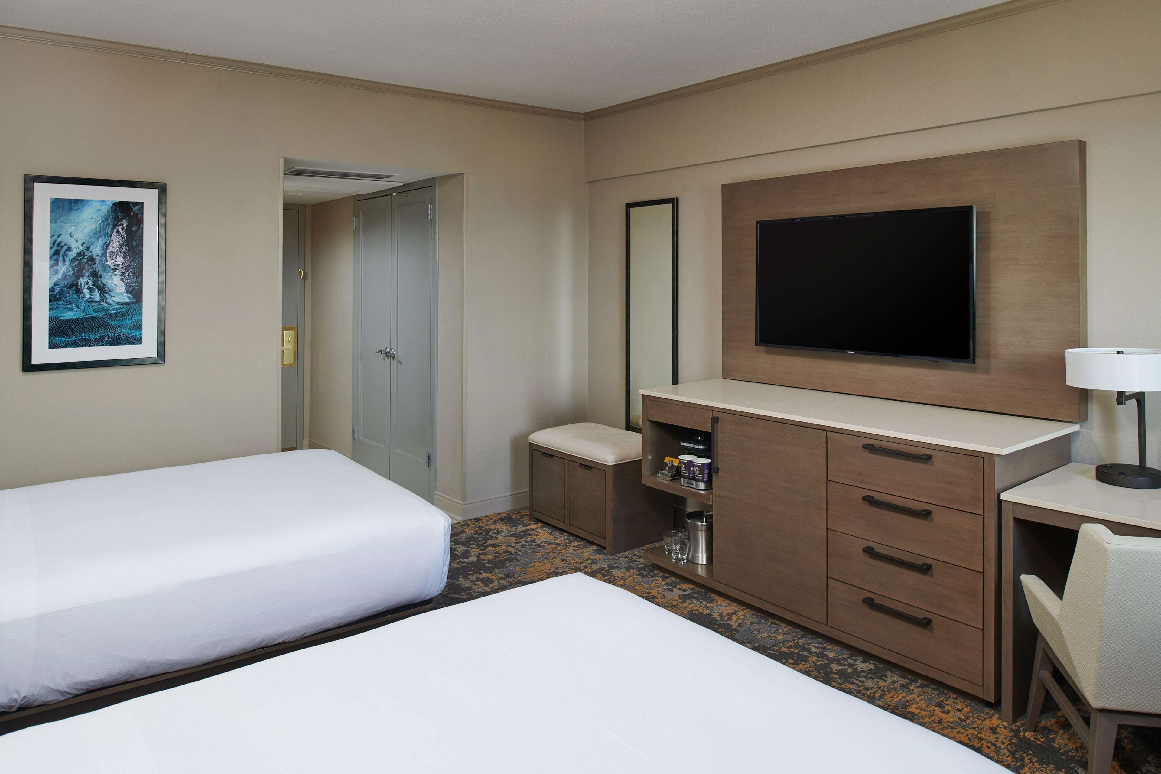 A hotel room with two beds, a TV, a desk, a chair, a small cabinet, and a framed picture on the wall.