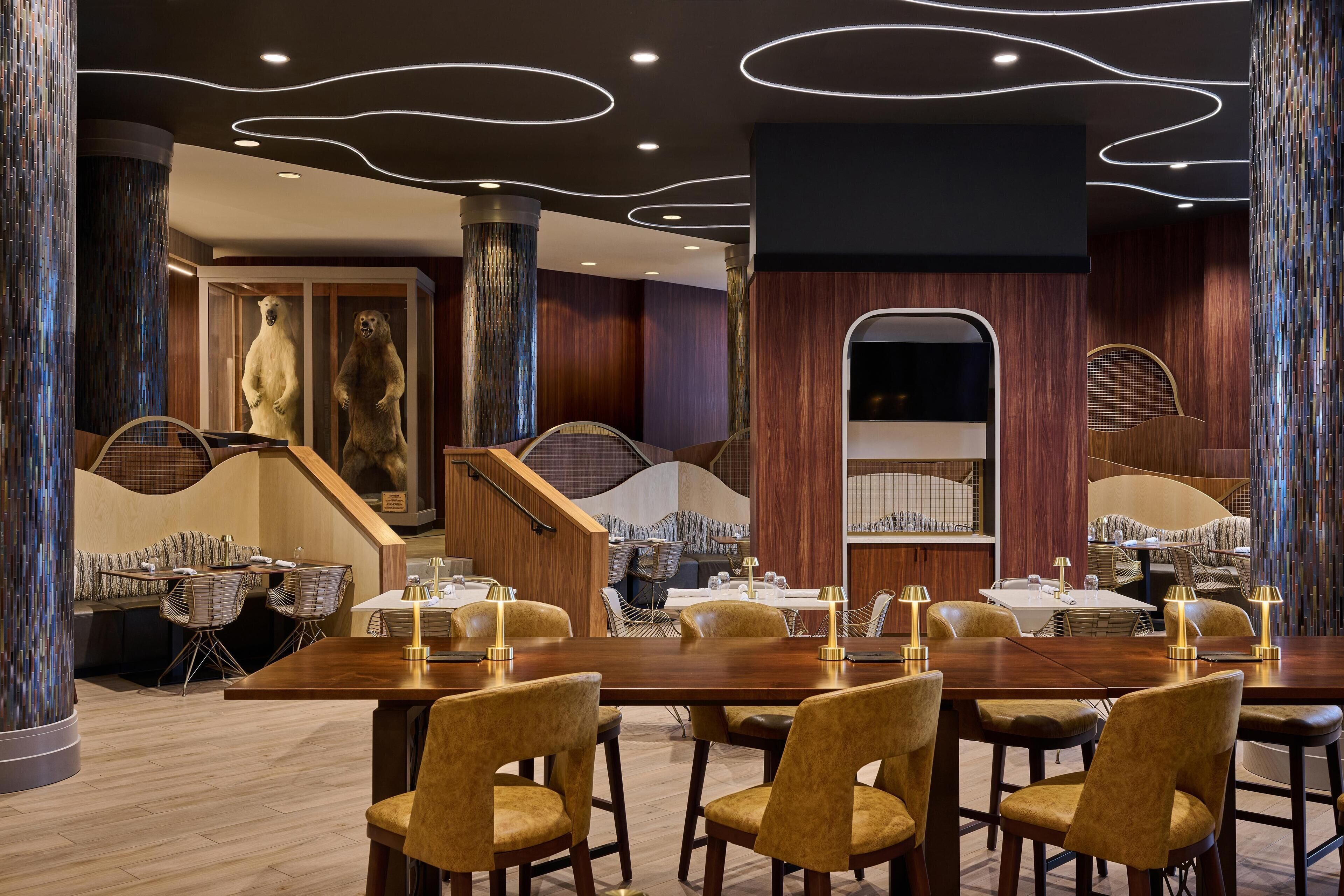 A modern dining area with wooden tables and chairs, a staircase, and decorative elements like a bear statue and a wall with a unique pattern.