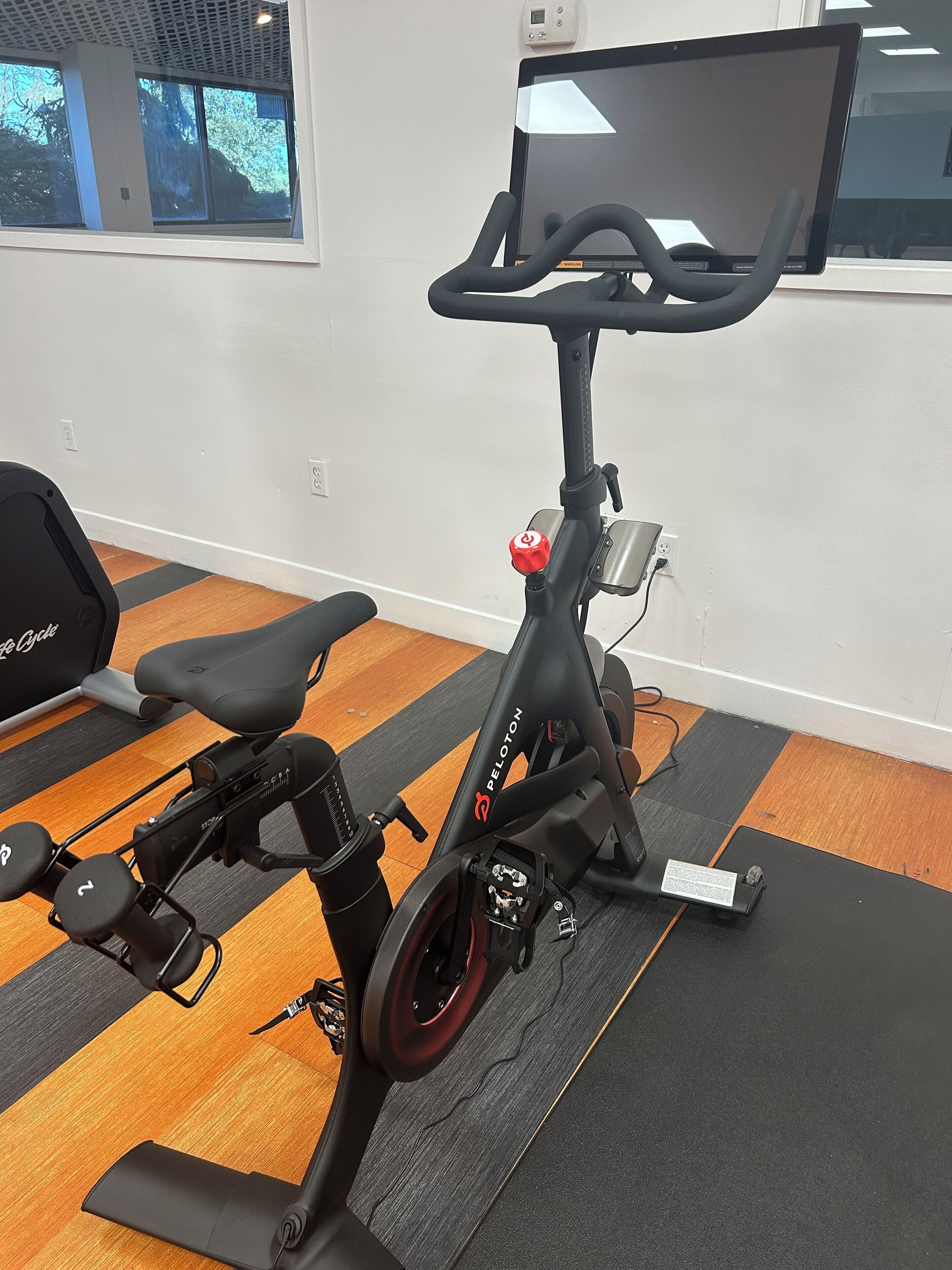 A Peloton stationary bike in a room with a large window, a treadmill, and a wall-mounted TV.