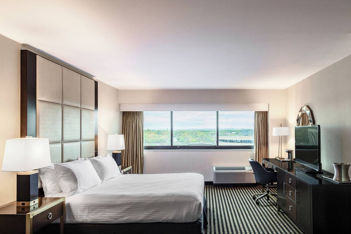 A hotel room with a large bed, two bedside tables with lamps, a desk with a chair, a flat-screen TV, and a window with a view of greenery.