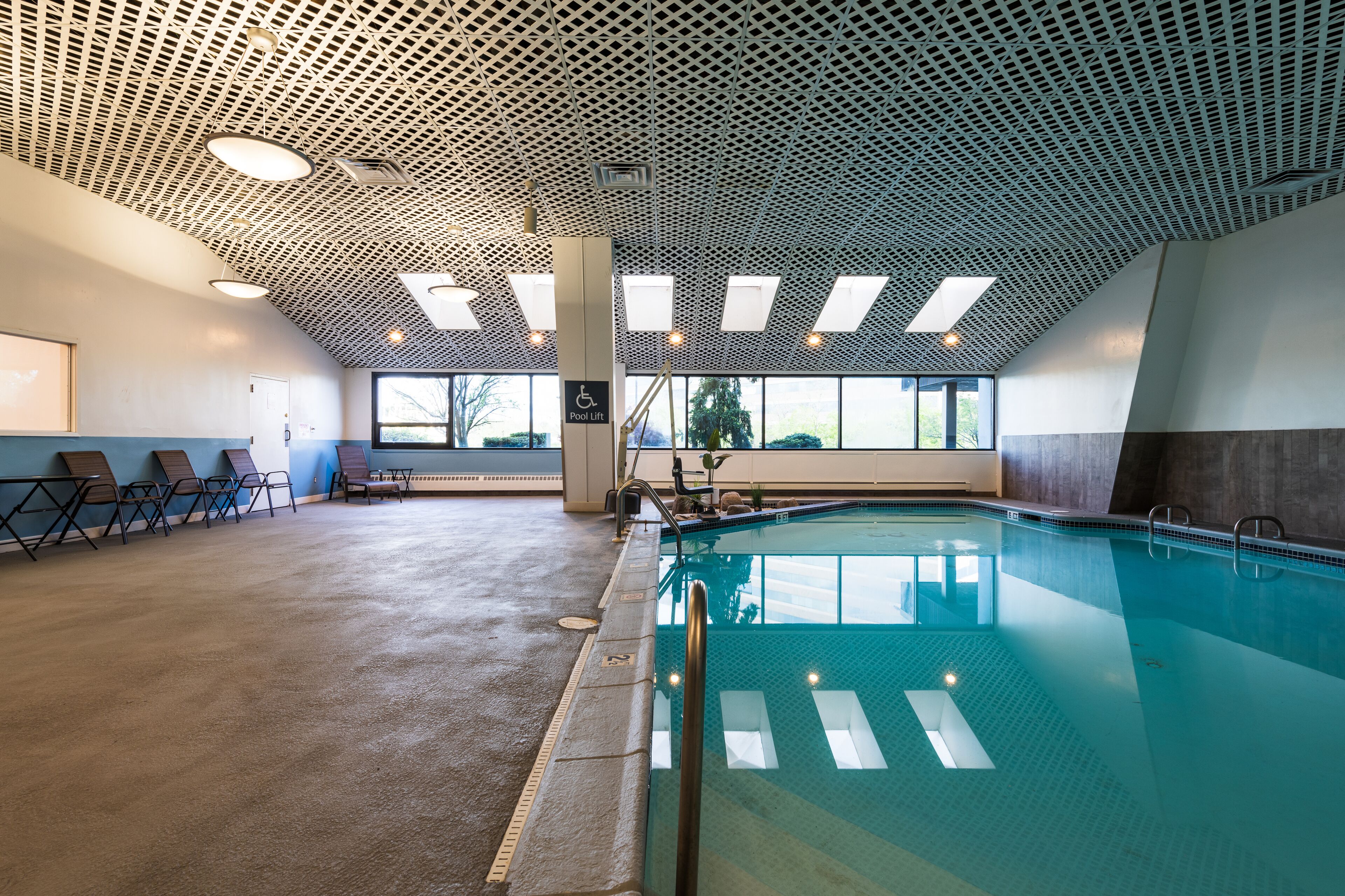 Piscina interior com teto em padrão geométrico, elevador de piscina e uma fileira de espreguiçadeiras ao longo da parede.