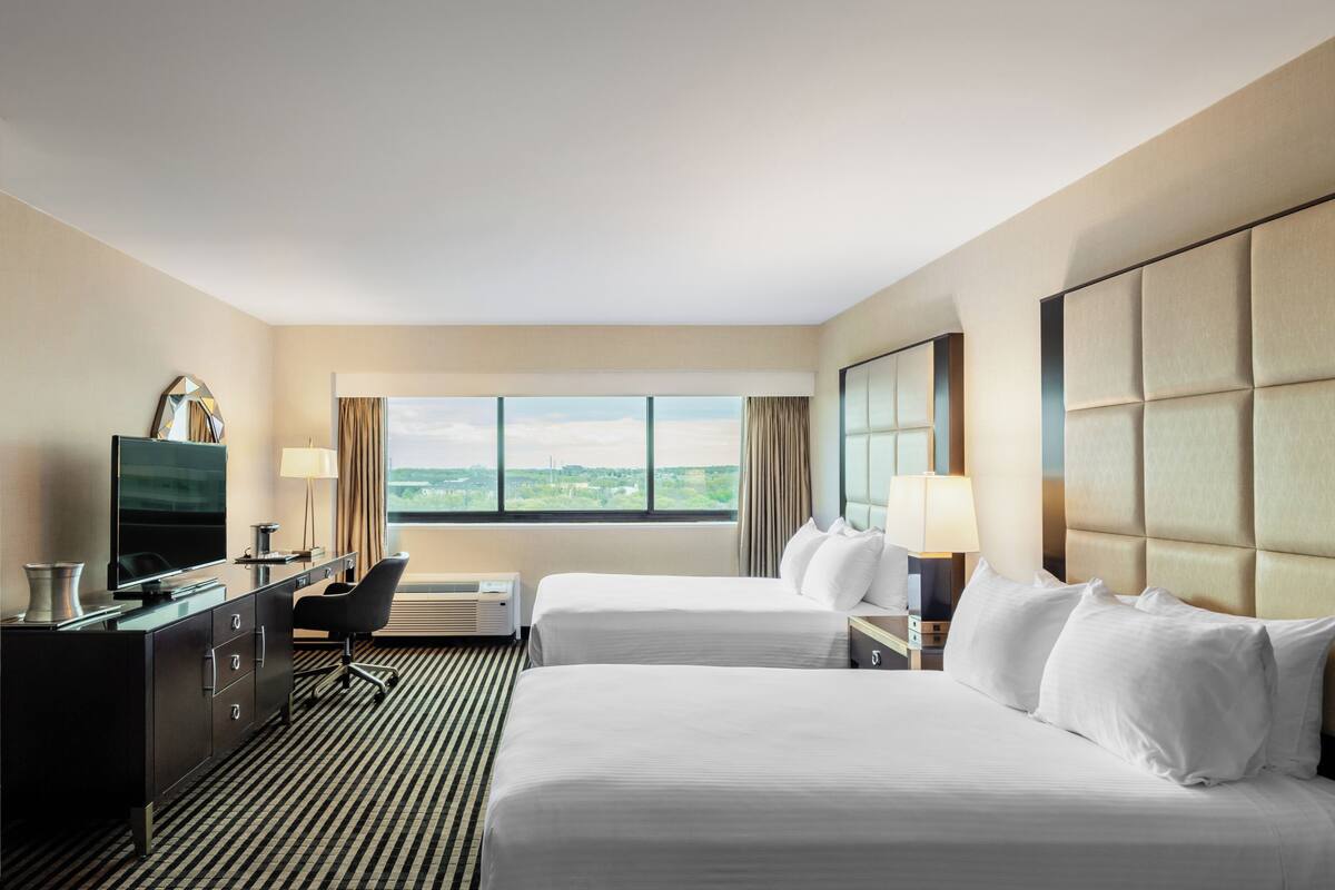 A hotel room with two beds, a desk with a TV, a chair, and a large window with a view.