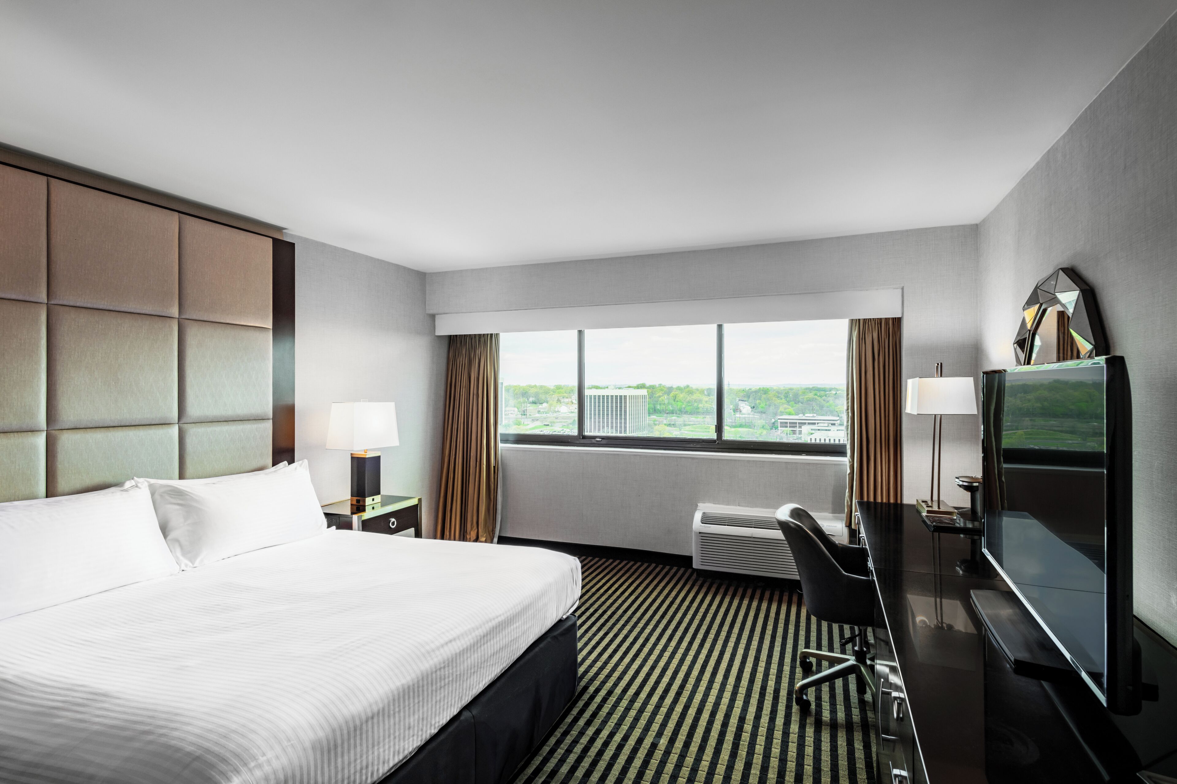 A hotel room with a large bed, a desk with a chair, a television, and a window with a city view.