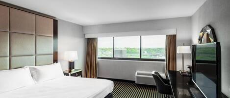 A hotel room with a large bed, a desk with a chair, a television, and a window with a city view.