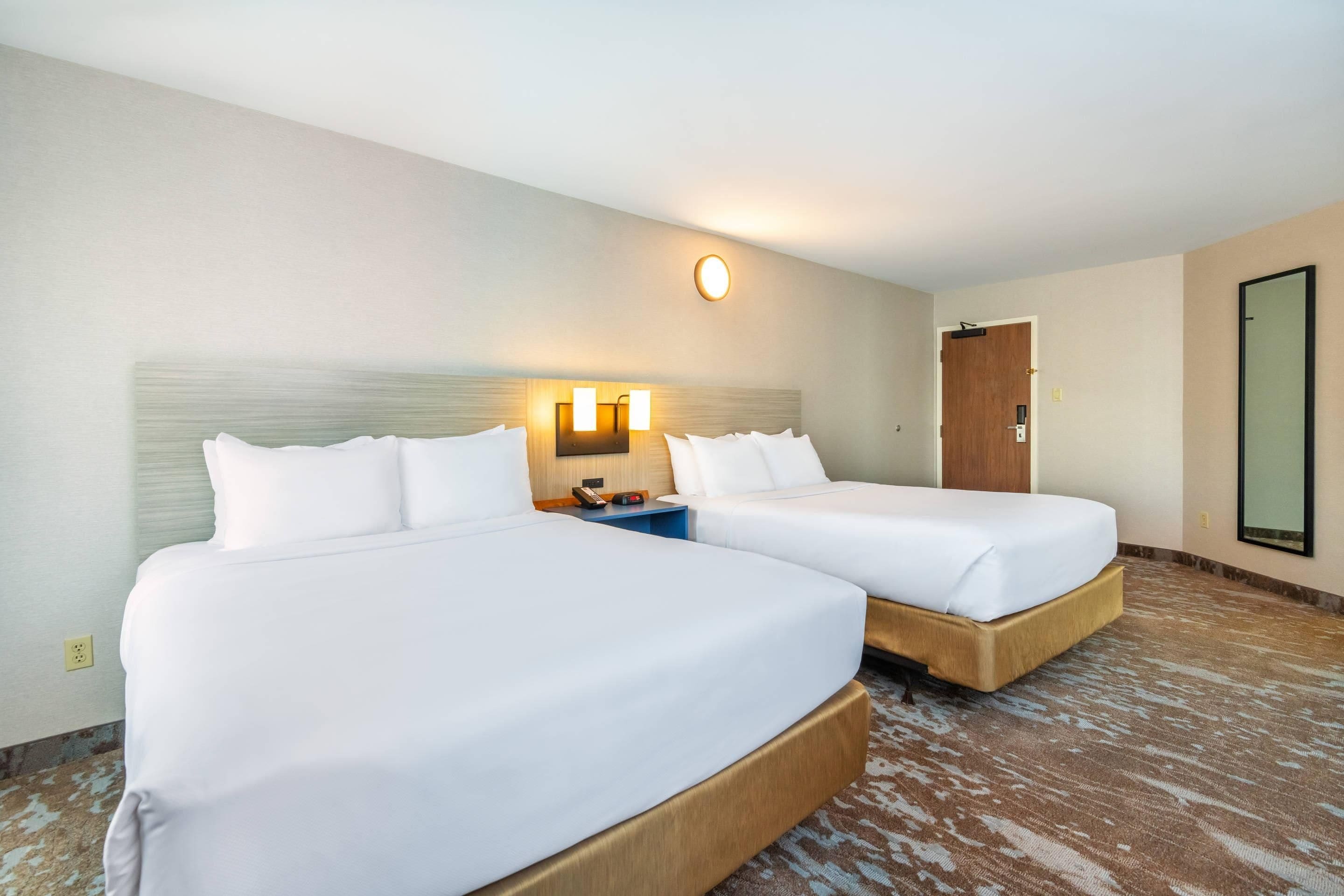 A hotel room with two beds, a nightstand, a wall-mounted light, and a door.