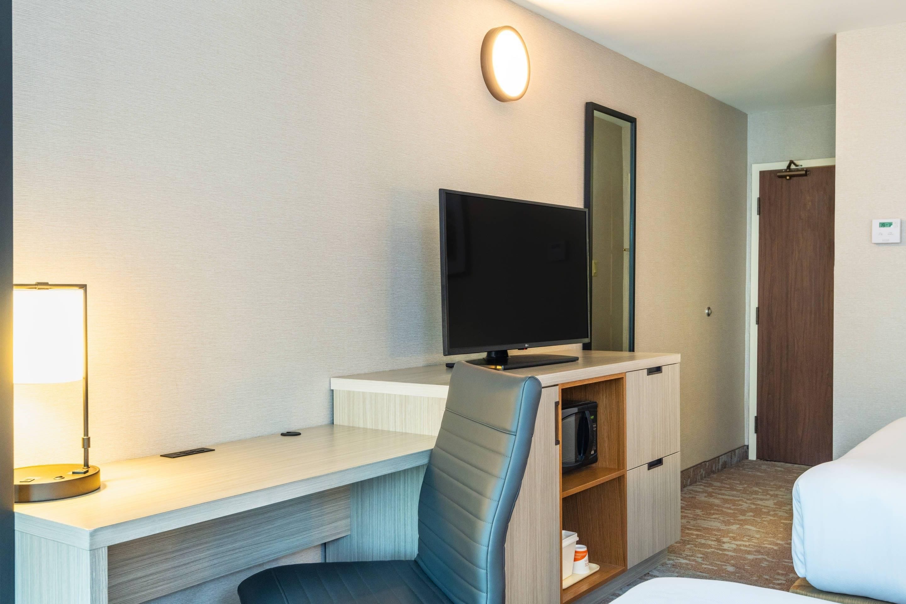 Ein Hotelzimmer mit Schreibtisch, Stuhl, Fernseher, Spiegel und Bett.