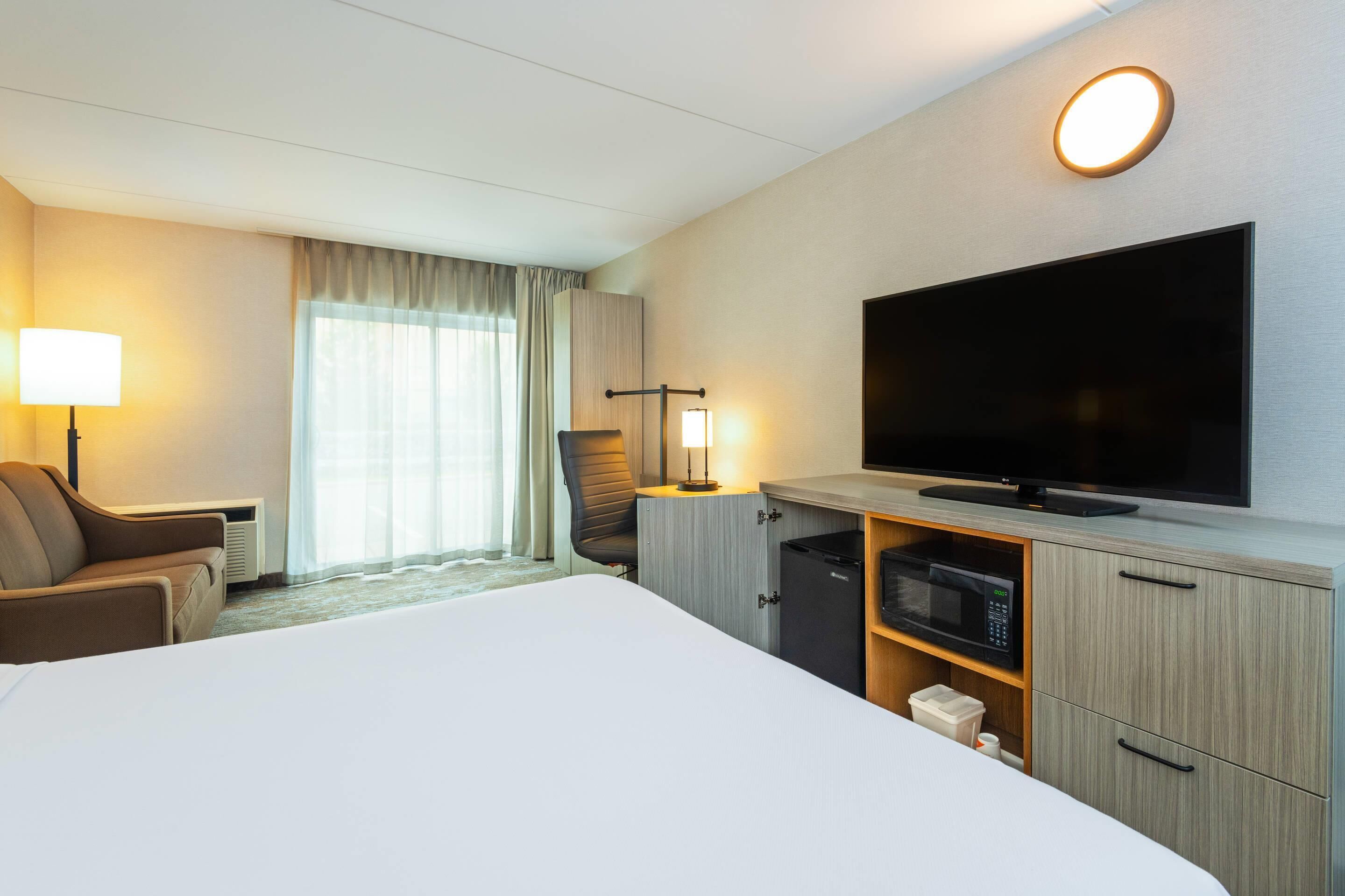 Habitación de hotel con una cama grande, televisión de pantalla plana, escritorio con lámpara, una silla y ventana con cortinas.