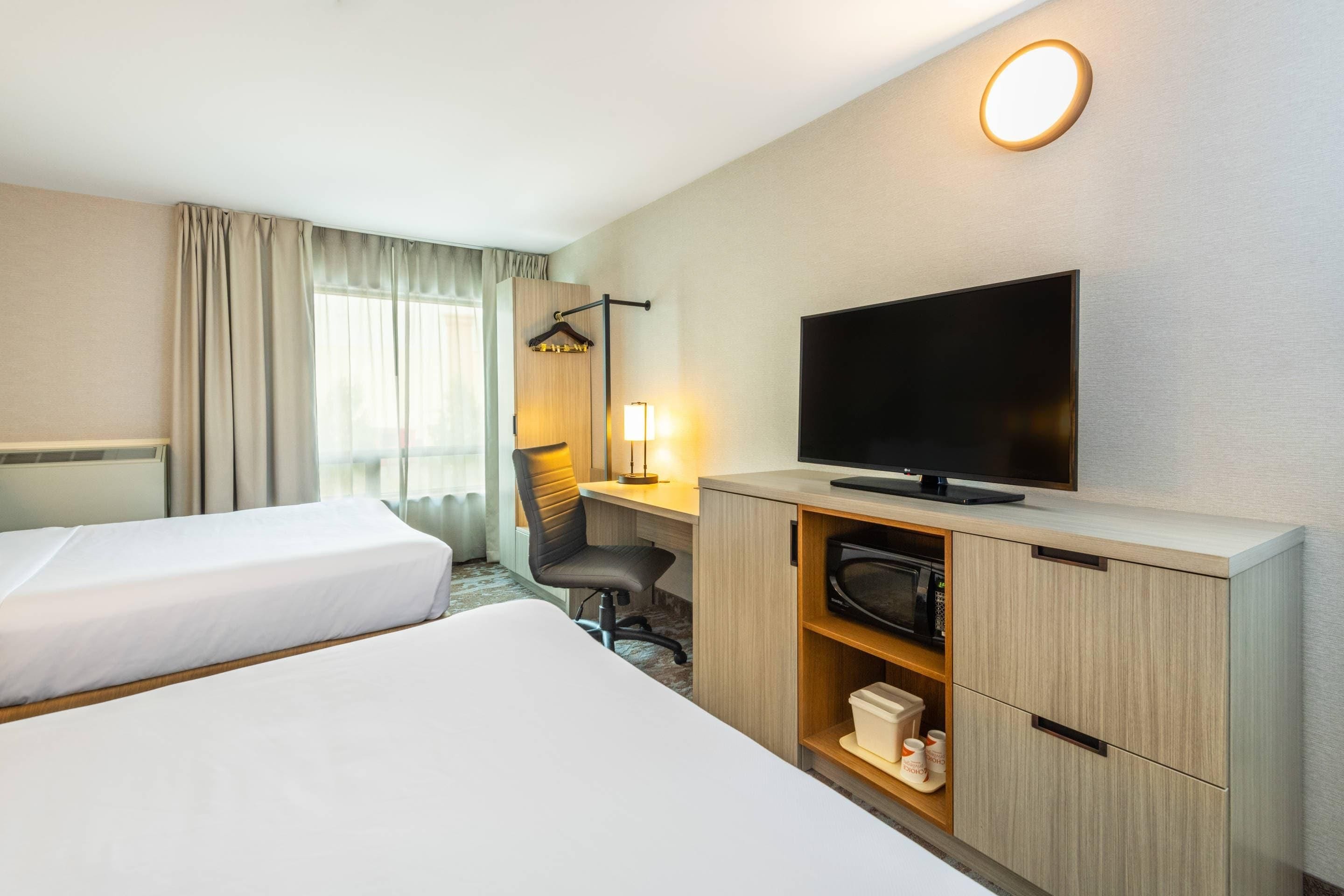 A hotel room with two beds, a desk with a flat-screen TV, a chair, a lamp, and a wardrobe.