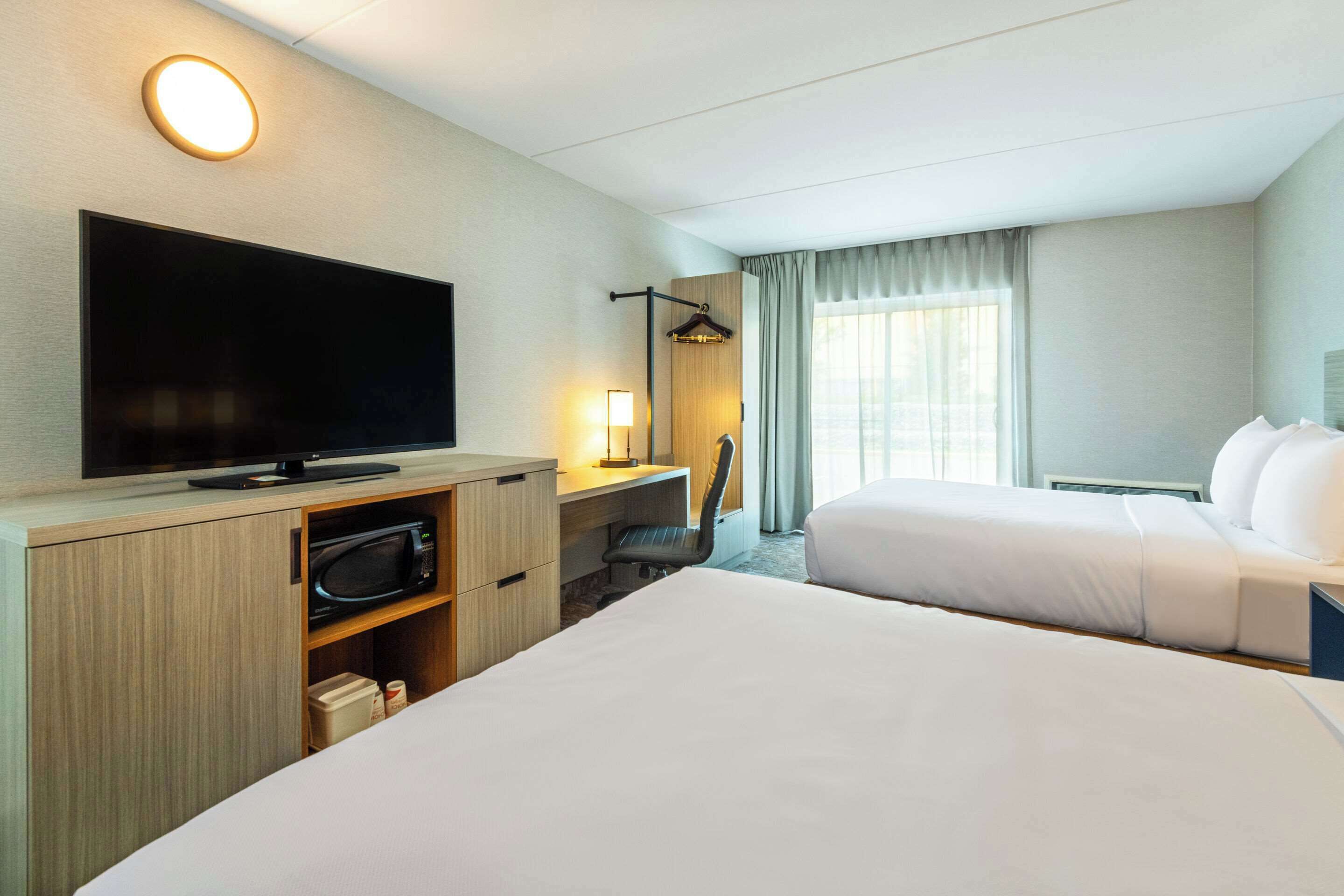 A hotel room with two beds, a flat-screen TV, a desk with a chair, a lamp, and a large window with curtains.