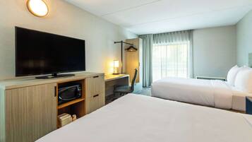 A hotel room with two beds, a flat-screen TV, a desk with a chair, a lamp, and a large window with curtains.