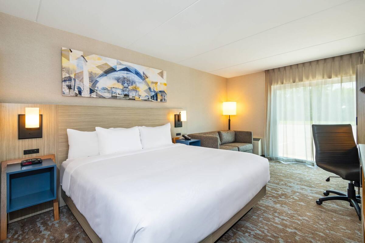 A hotel room with a large bed, a desk with a chair, two bedside tables with lamps, and a large window with curtains.