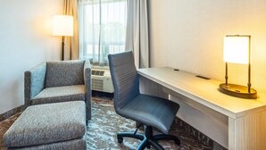 Habitación de hotel con escritorio, dos sillones, una lámpara de pie y un ventanal con cortinas.
