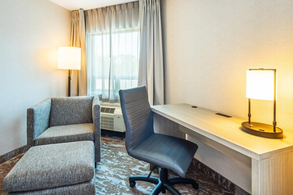 Habitación de hotel con escritorio, dos sillones, una lámpara de pie y un ventanal con cortinas.