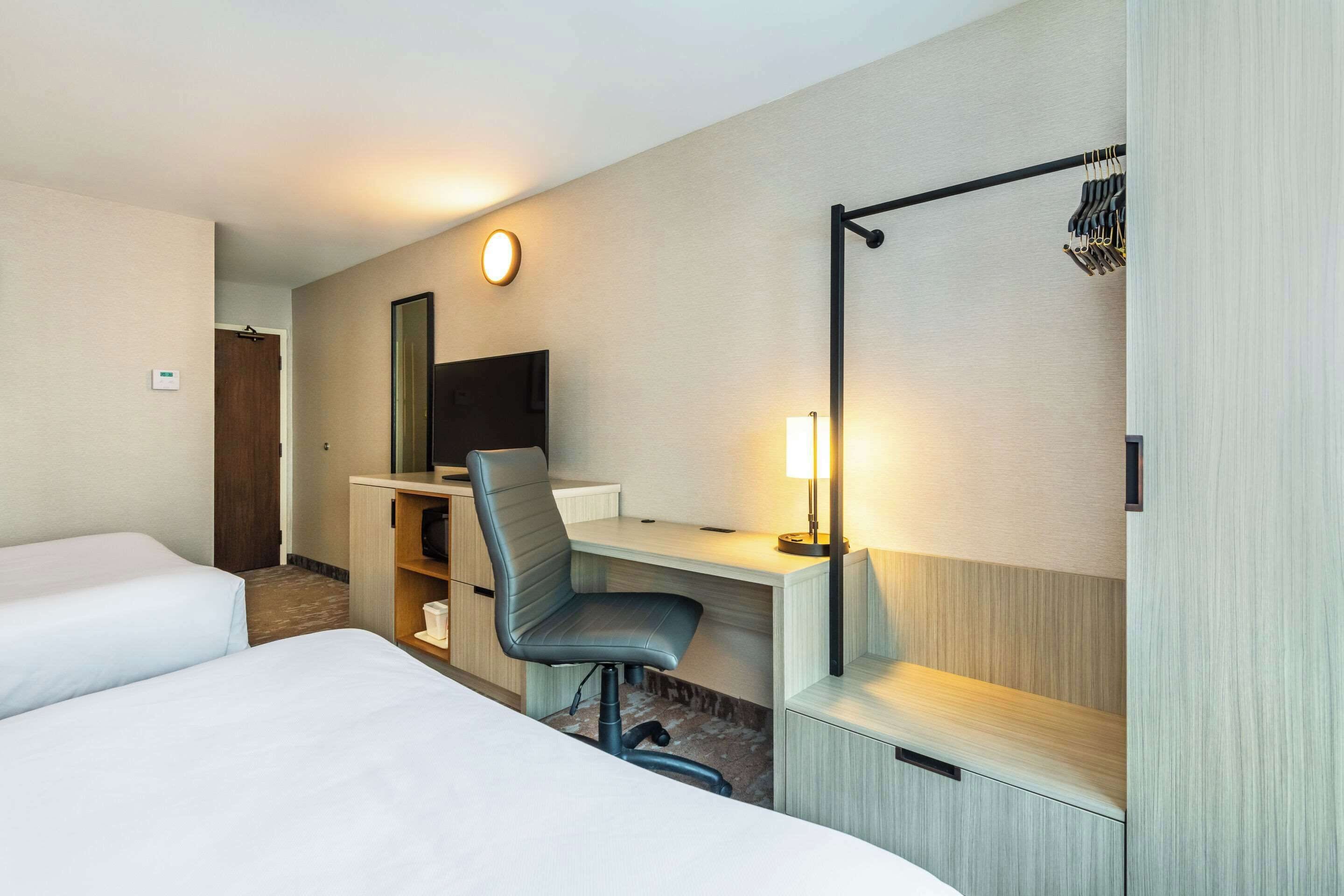 A hotel room with two beds, a desk with a chair, a TV, and a lamp.