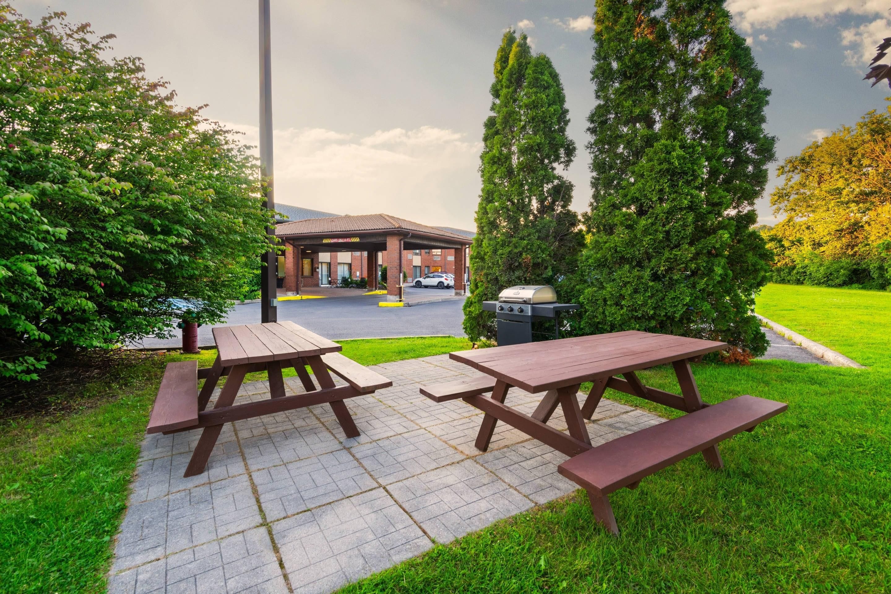 Un área de picnic con dos mesas y bancos de madera, una parrilla y un estacionamiento al fondo.