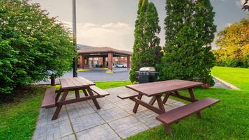 A picnic area with two wooden tables and benches, a barbecue grill, and a parking lot in the background.