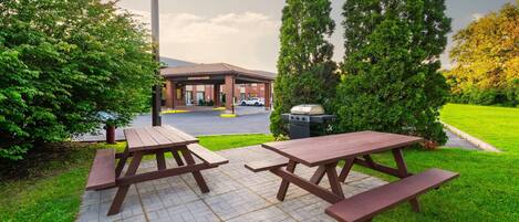 Un área de picnic con dos mesas y bancos de madera, una parrilla y un estacionamiento al fondo.