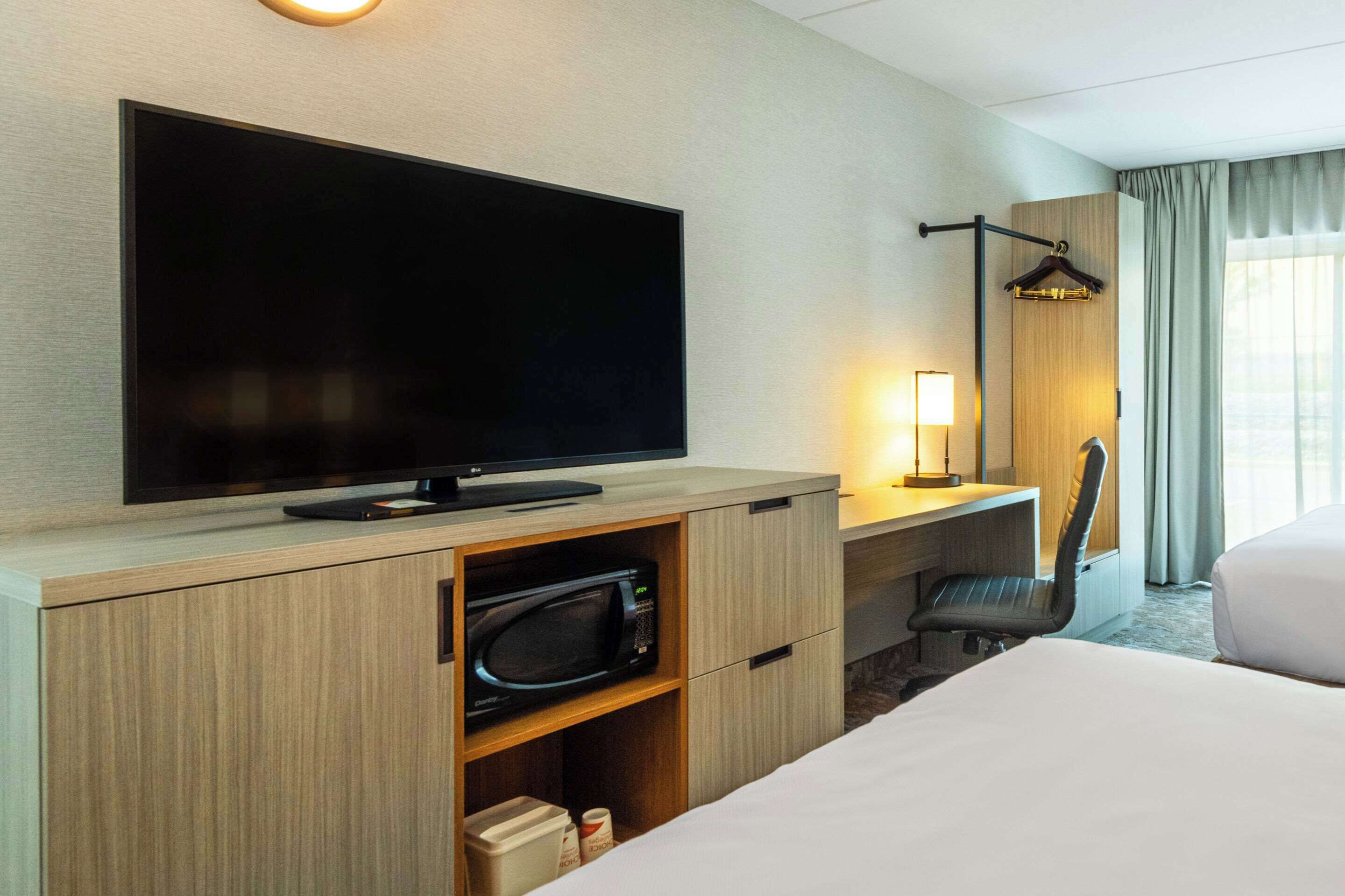 A hotel room with a large flat-screen TV, a desk with a lamp, a microwave, and a chair.