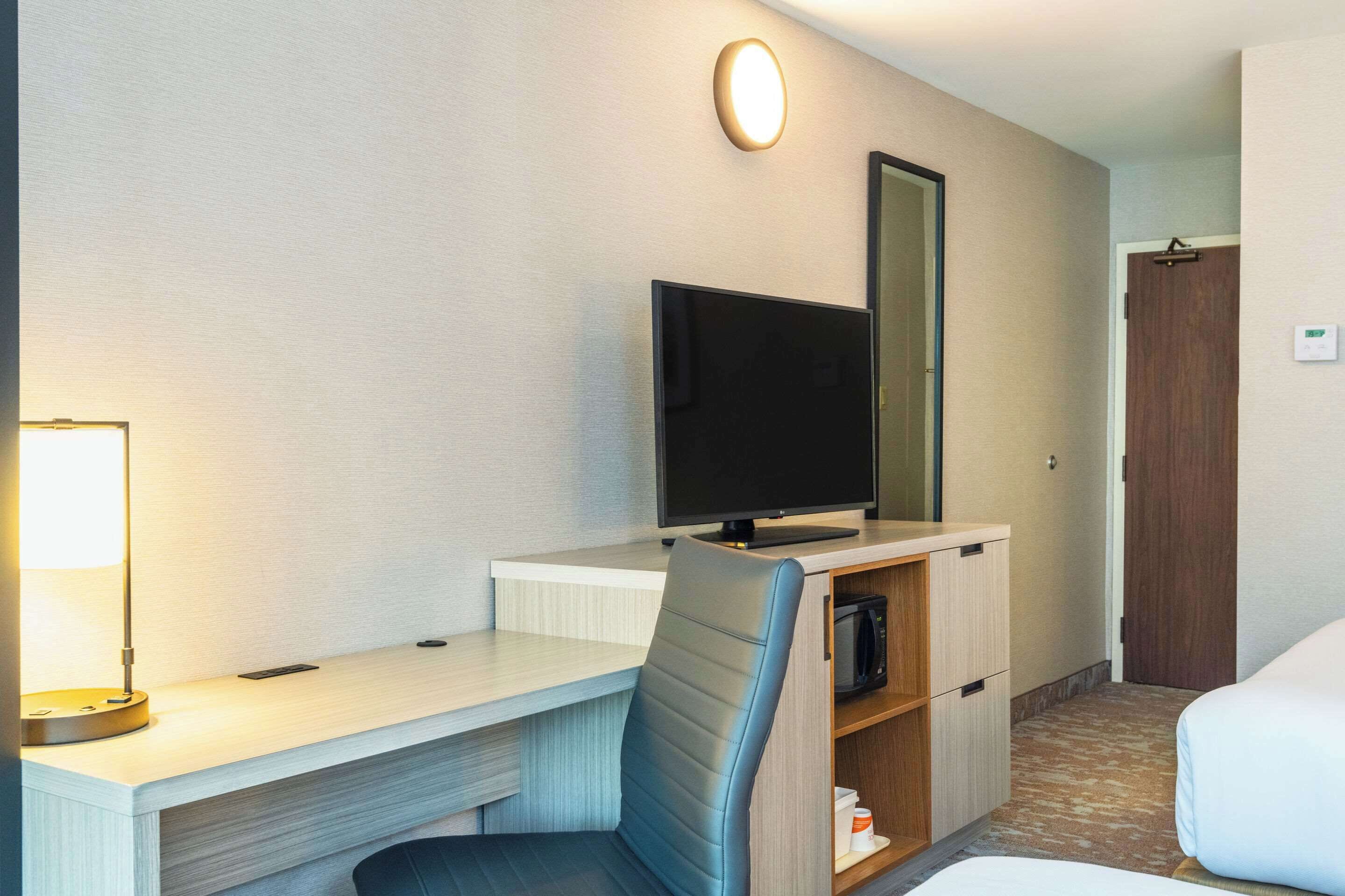 A hotel room with a desk, a flat-screen TV, a chair, a bed, and a door.