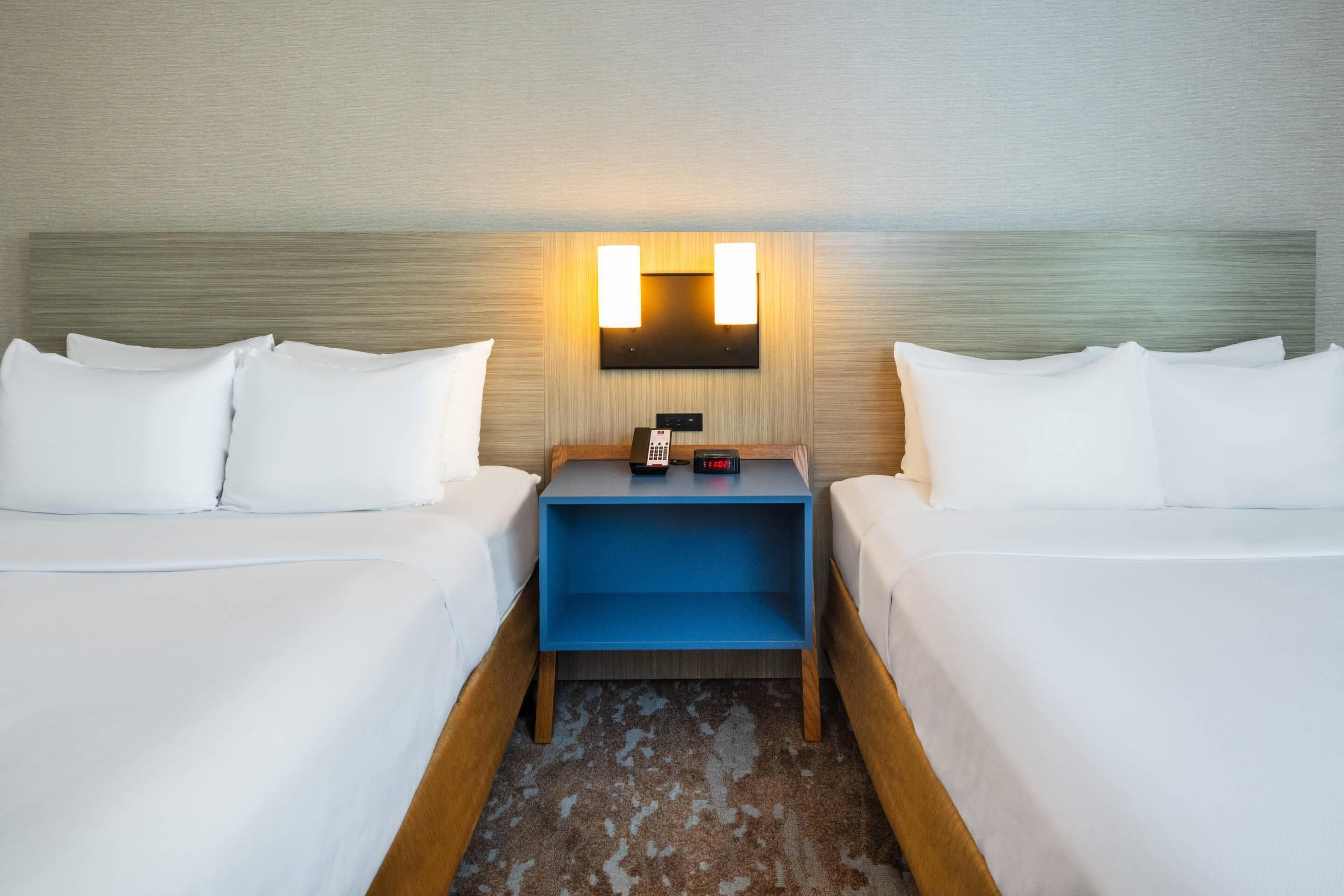 A hotel room with two beds, a nightstand with a clock, and a wall-mounted light fixture.