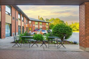 Un patio al aire libre con mesas y sillas, un edificio de ladrillo al fondo y un estacionamiento con coches.