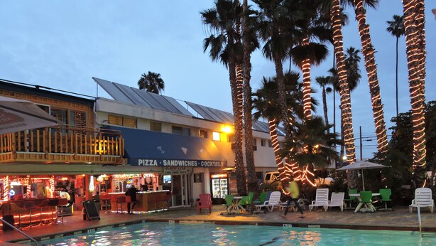 A pool area with lounge chairs and a bar, surrounded by palm trees and a building with a sign for pizza and sandwiches. - Los Angeles Adventurer All Suite Hotel at LAX (Inglewood)