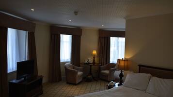 Ein Hotelzimmer mit einem Bett, zwei Sesseln, einem kleinen Tisch mit einer Lampe, einem Fernseher und Vorhängen an den Fenstern.