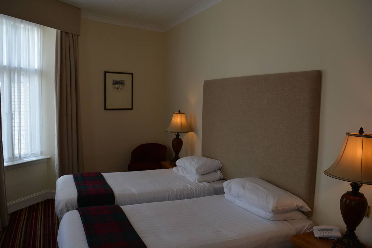 A hotel room with two beds, a nightstand with a lamp, a chair, and a framed picture on the wall.
