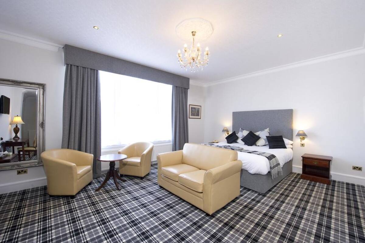 A hotel room with a bed, two armchairs, a small table, a mirror, and a chandelier.