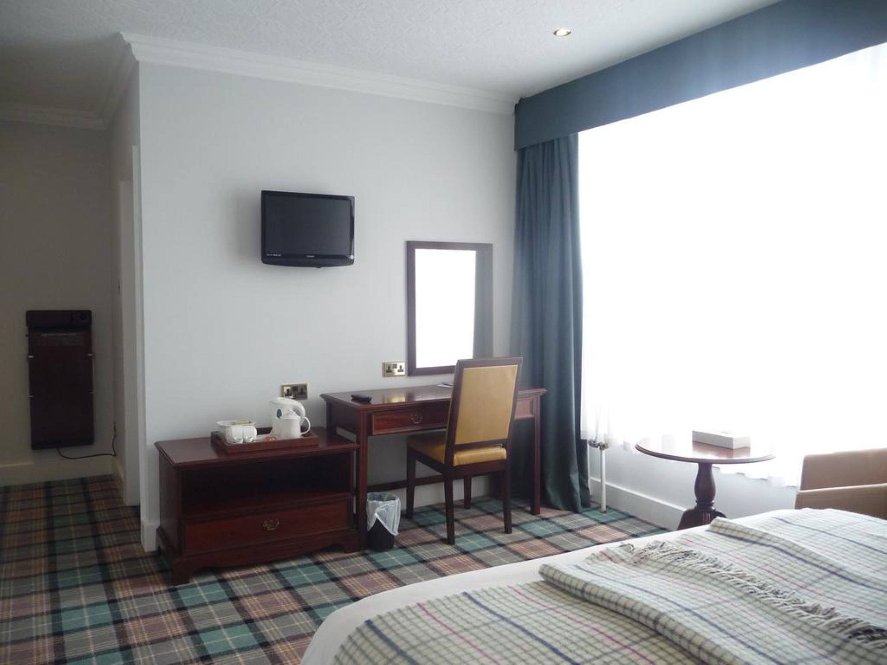A hotel room with a bed, a desk with a chair, a television mounted on the wall, a window with curtains, and a small table.