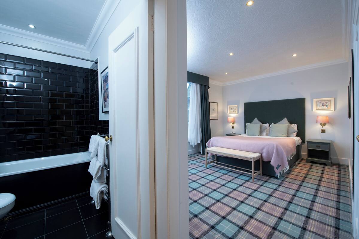 A hotel room with a black-tiled bathroom, a bed with a plaid-patterned floor, and a bench.
