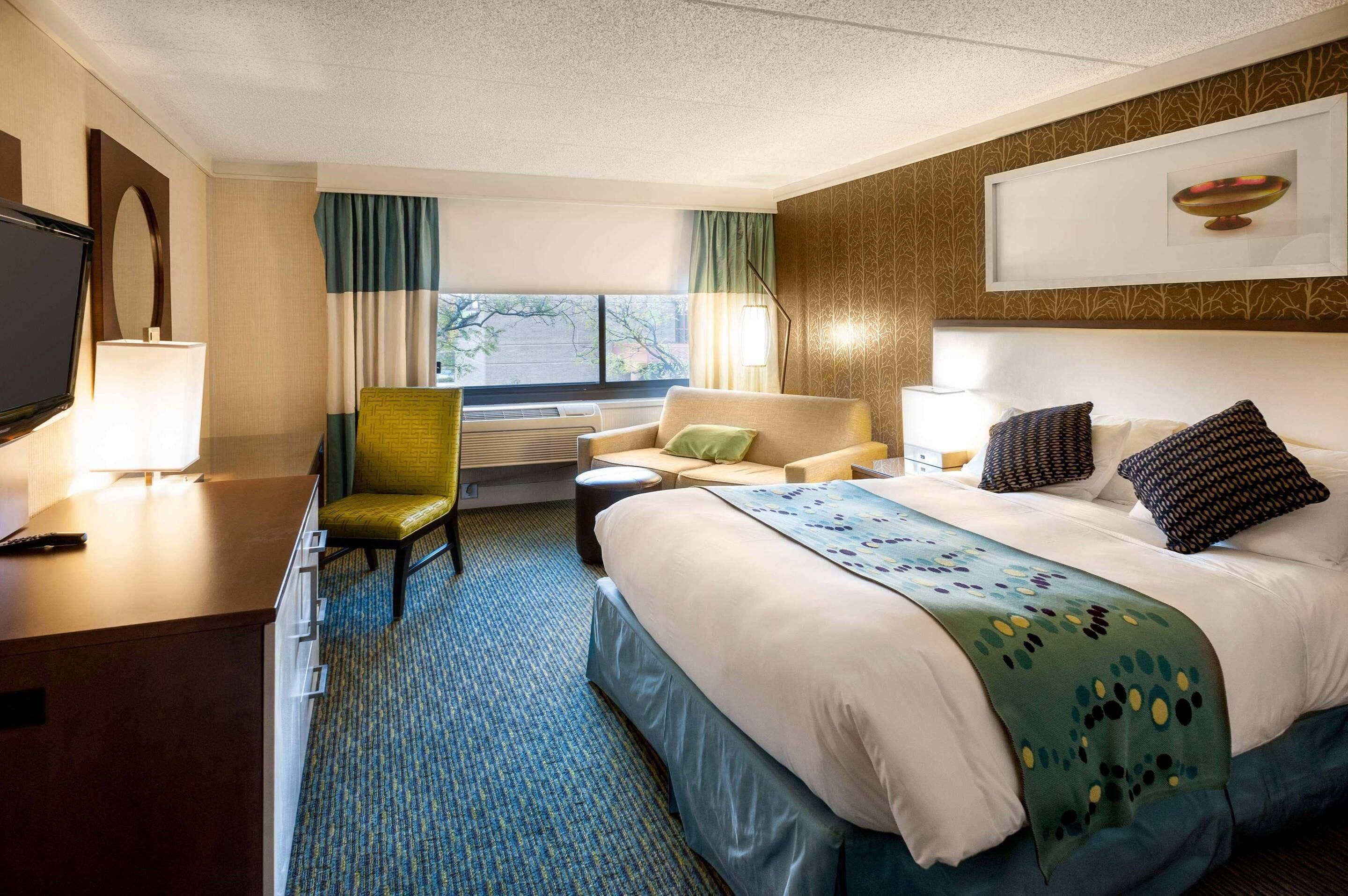 Une chambre d’hôtel avec un lit, un bureau, une chaise, une télévision et une fenêtre avec des rideaux.