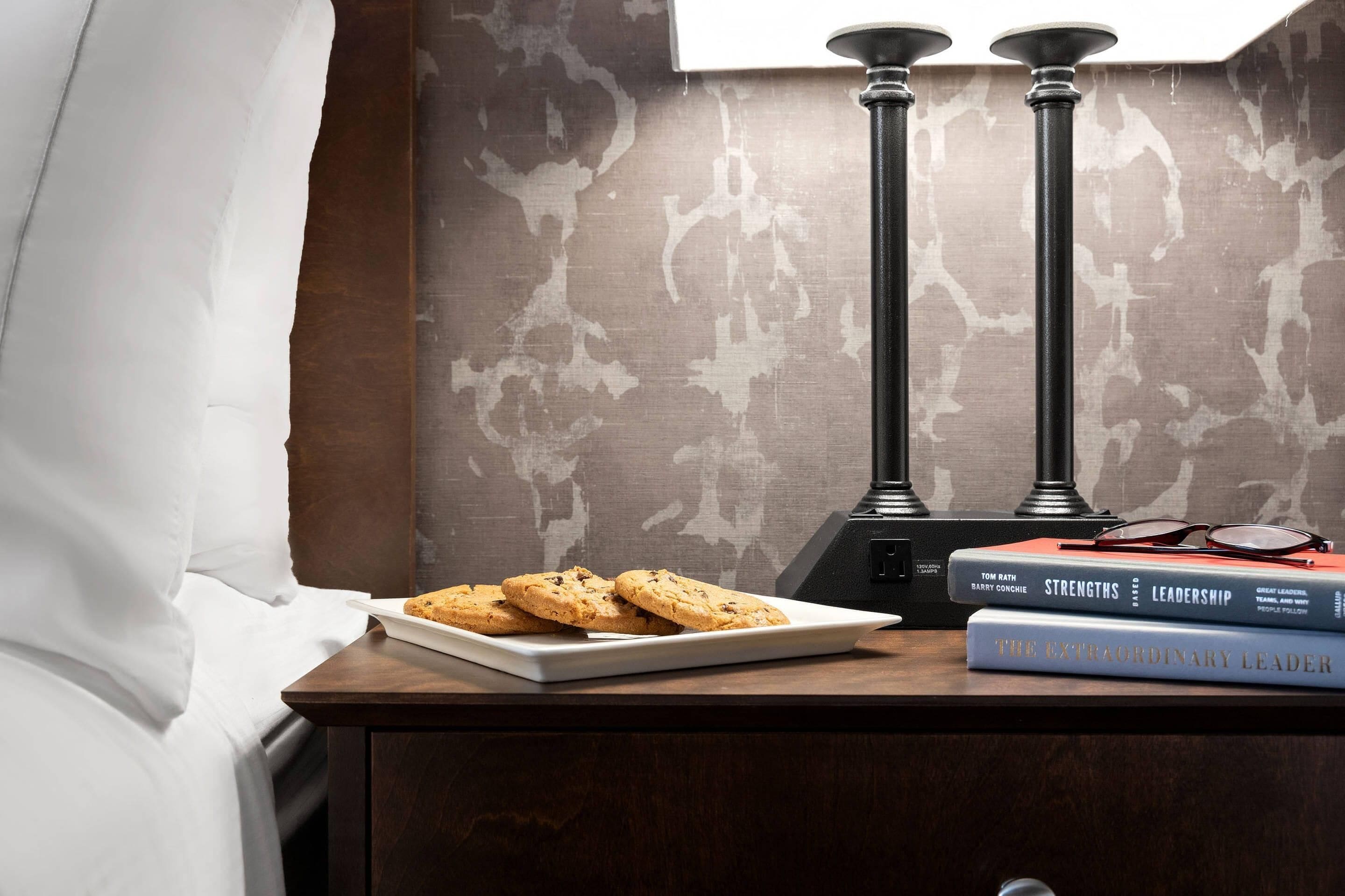 A neatly made bed with white linens, a plate of cookies, and two books titled 'Strengths of Leadership' and 'The Extraordinary Leader' on a wooden nightstand.