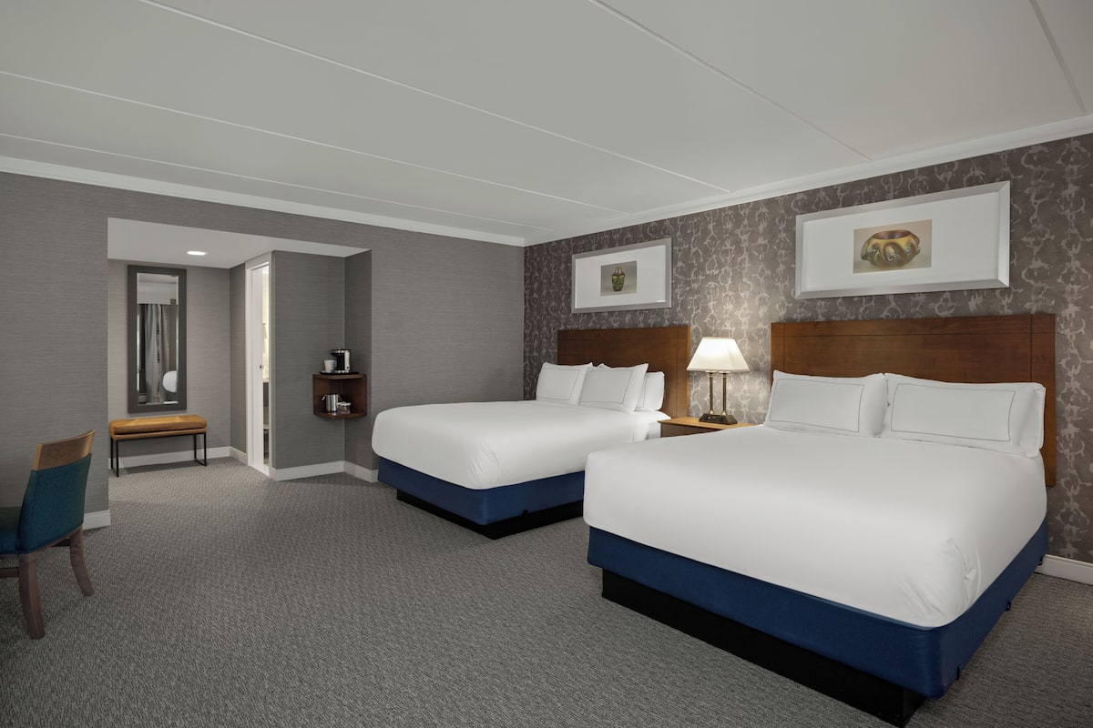 Une chambre d’hôtel avec deux lits, un banc, une chaise, une table de chevet et un tableau accroché au mur.