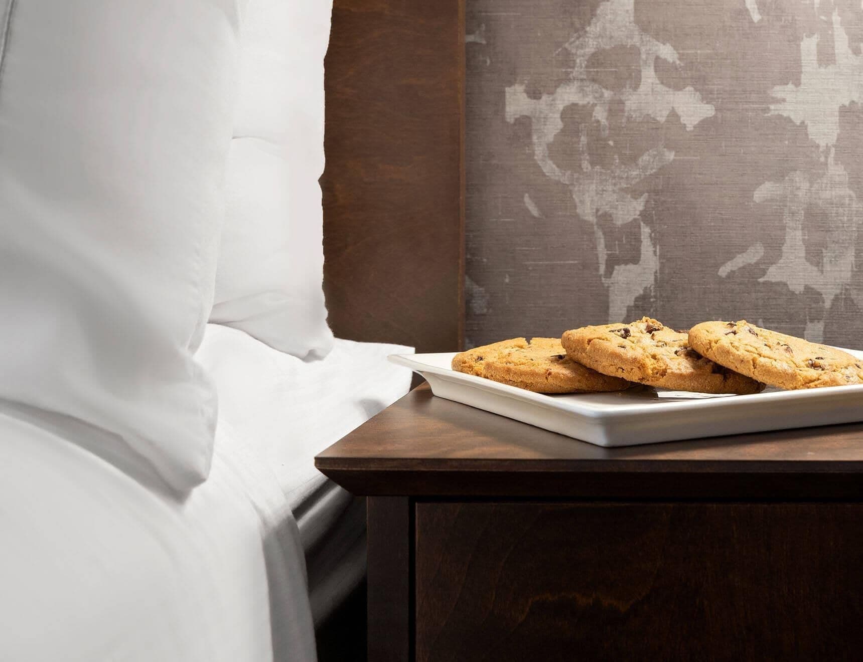 Uma cama com lençóis brancos, um criado-mudo de madeira com um prato de biscoitos e um mapa-múndi na parede.