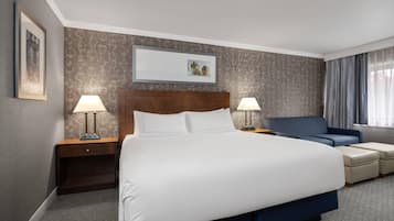 A hotel room with a large bed, two bedside tables with lamps, a blue armchair, a small table, and a framed picture on the wall.