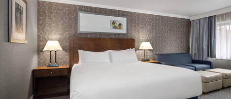 A hotel room with a large bed, two bedside tables with lamps, a blue armchair, a small table, and a framed picture on the wall.