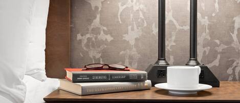 A bedside table with books, glasses, and a white cup on a wooden nightstand.