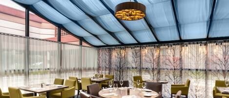 A restaurant with round tables and chairs, a large glass ceiling, and a view of trees through the windows.
