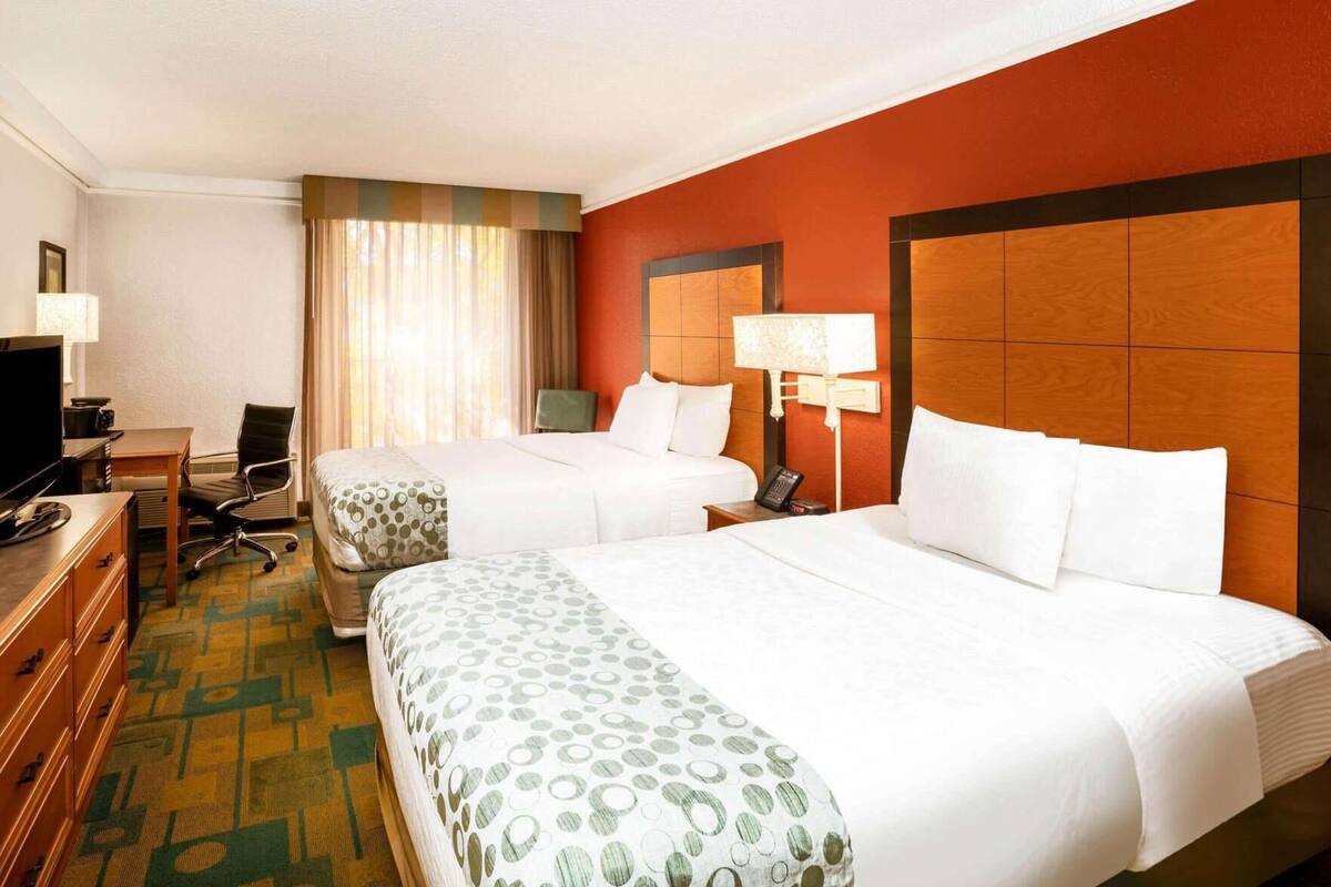Quarto de hotel com duas camas, uma escrivaninha com computador, uma cadeira, televisão e janela com cortinas.
