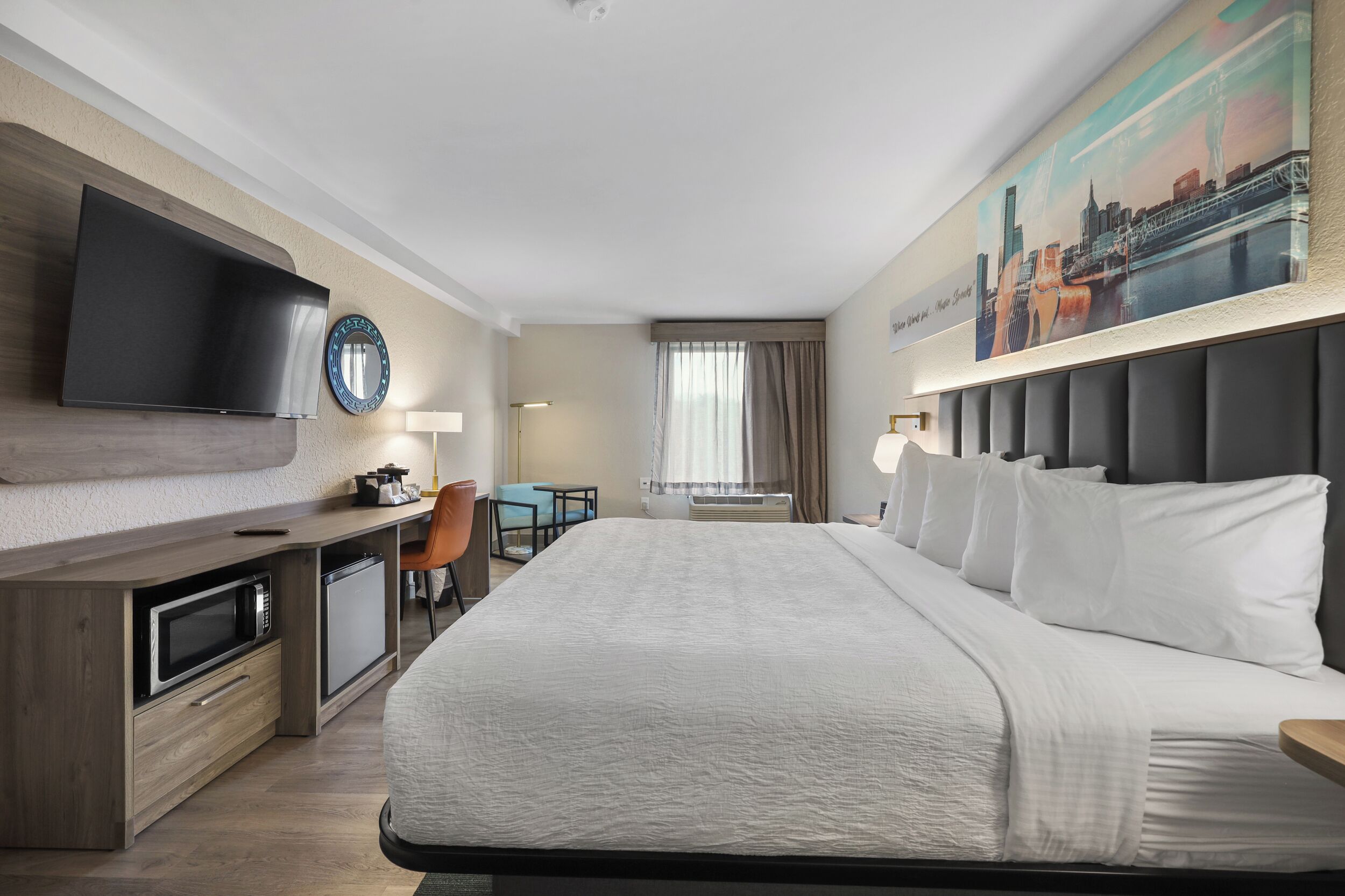 A hotel room with a bed, a desk with a microwave, a flat-screen TV, a chair, and a window with curtains.