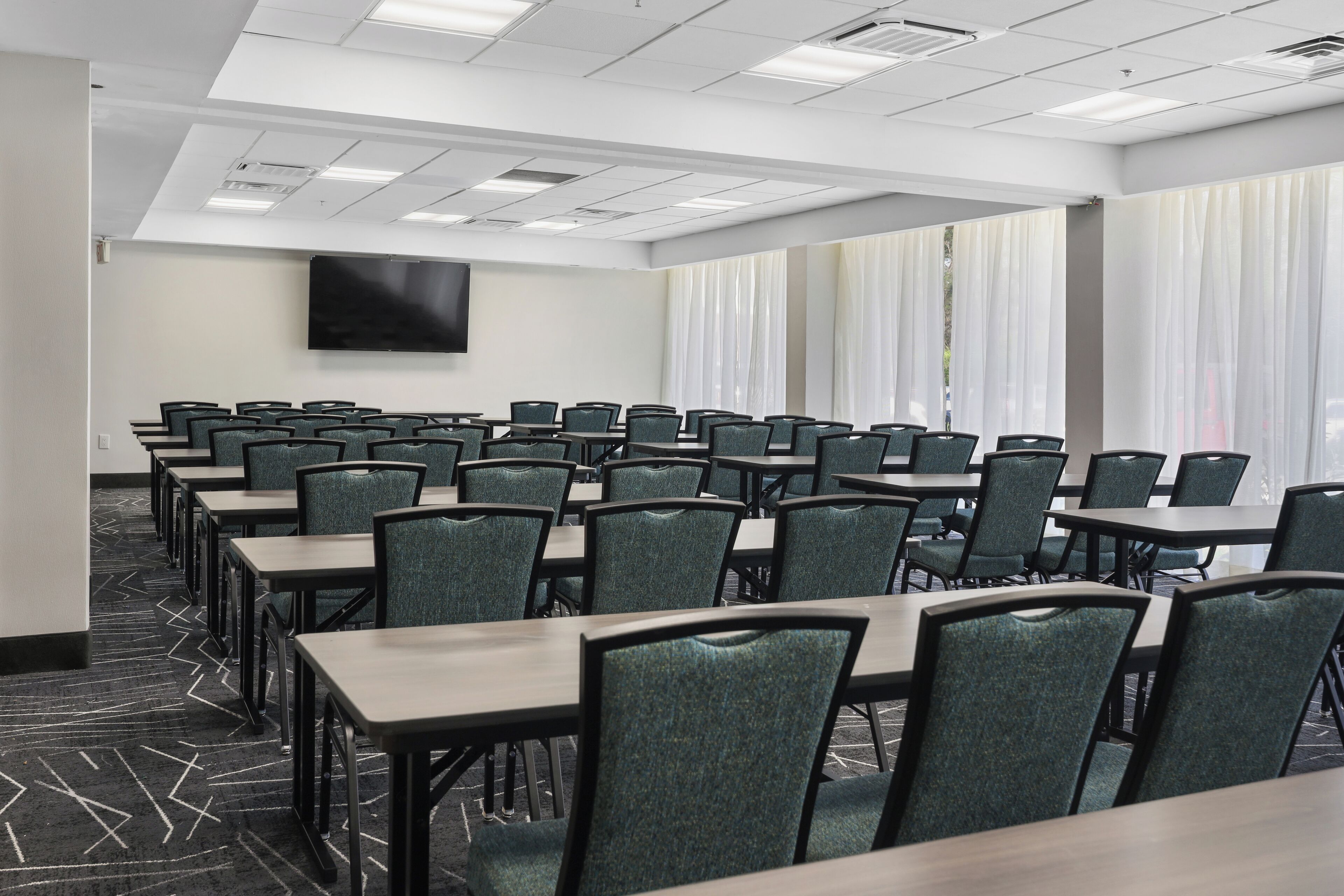 A conference room with rows of tables and chairs, a flat-screen TV on the wall, and large windows with curtains.