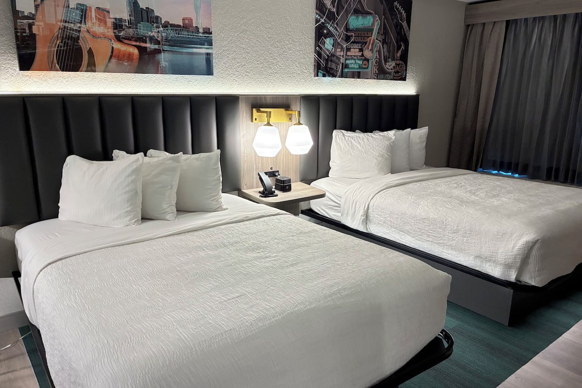 Quarto de hotel com duas camas, uma mesa de cabeceira com abajur e uma obra de arte na parede.