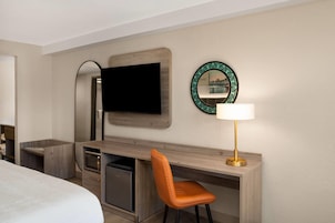 A hotel room with a bed, a desk with a chair, a television, a mirror, and a lamp.