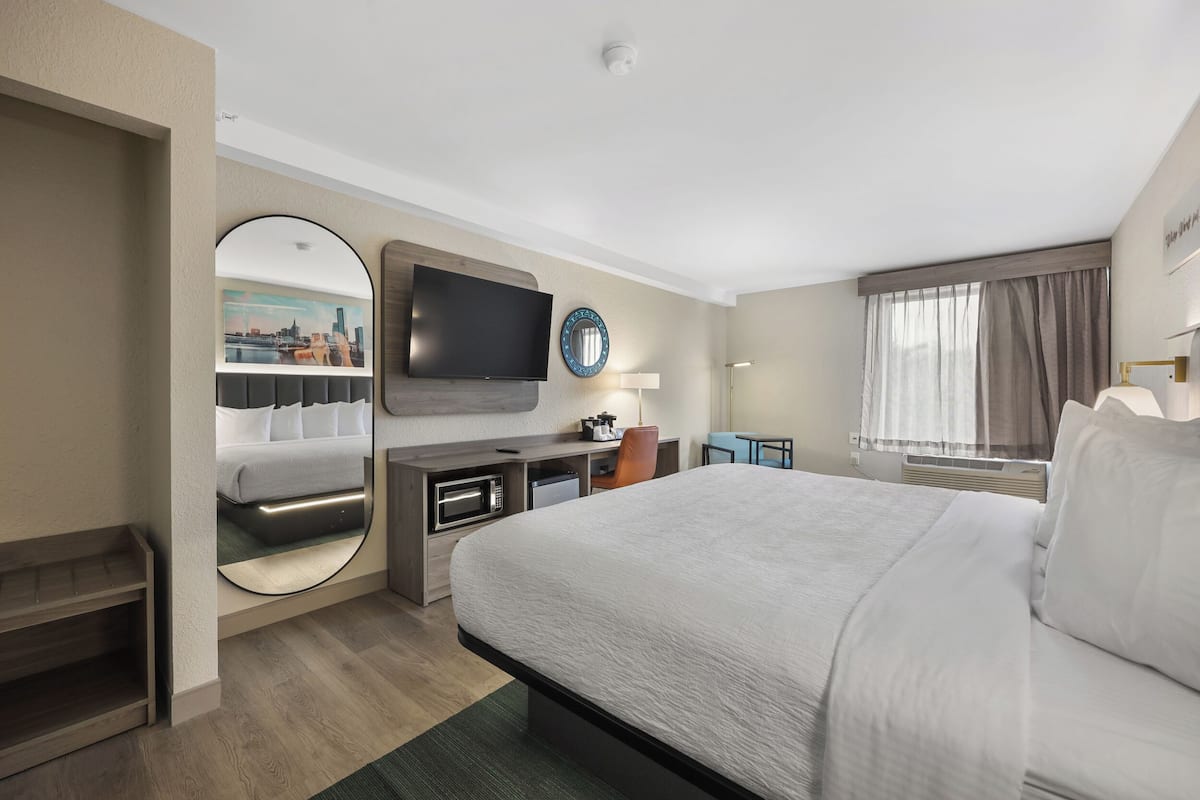 Quarto de hotel com uma cama grande, televisão de tela plana, escrivaninha com cadeira, espelho redondo e janela com cortinas.