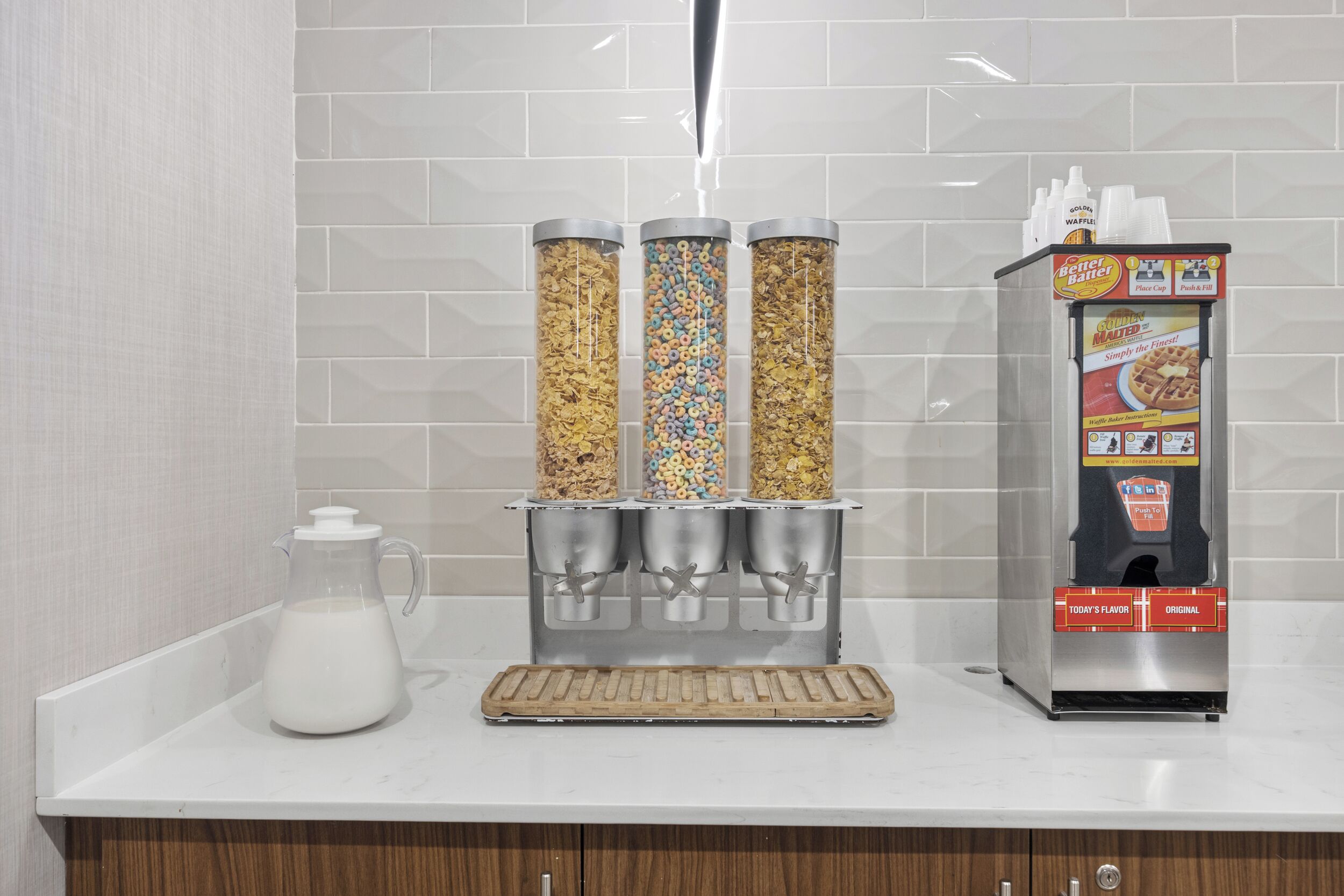 A cereal dispenser with three containers, a milk pitcher, and a waffle machine on a countertop.
