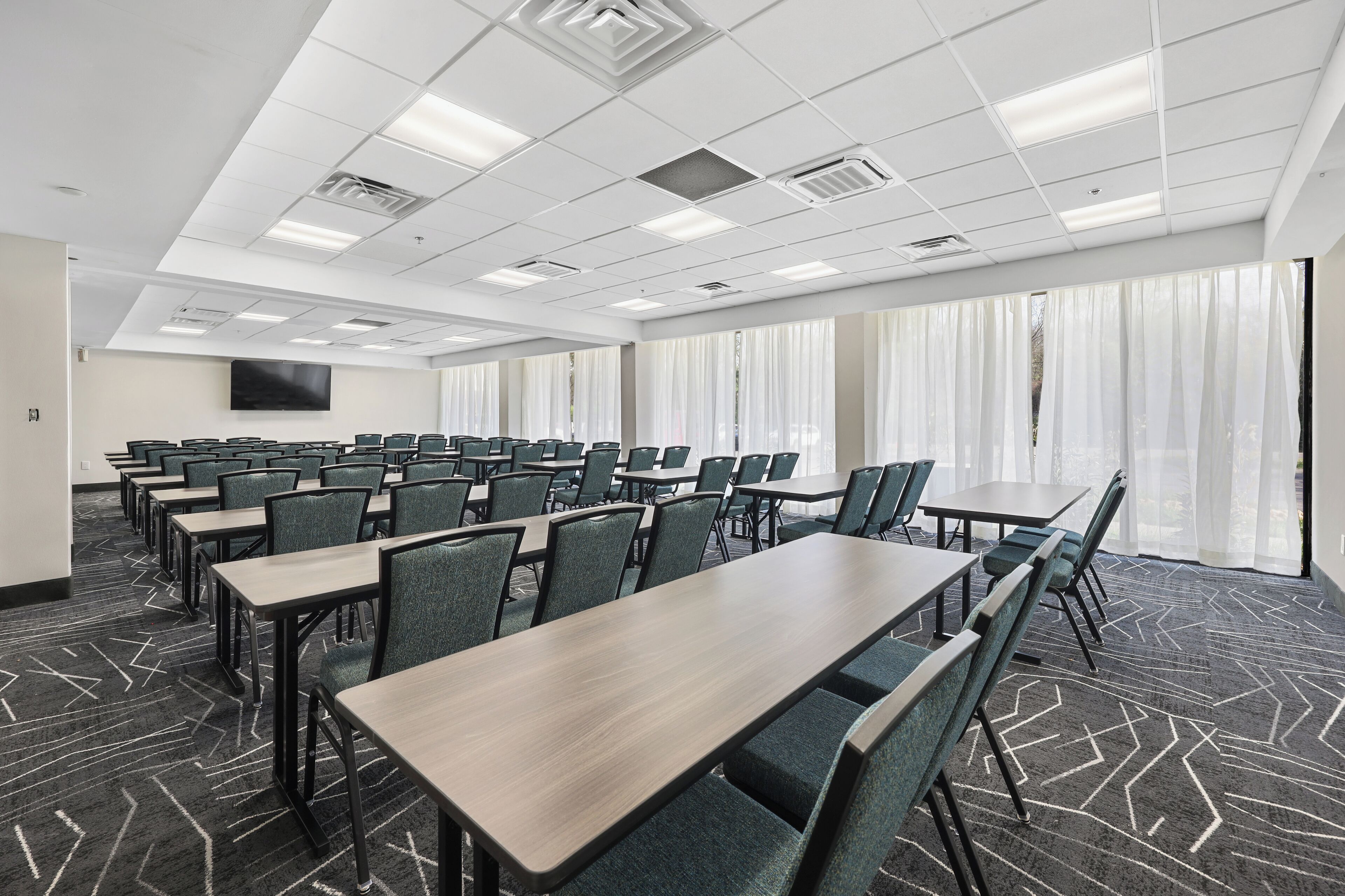 Un salón de conferencias con filas de mesas y sillas, un televisor de pantalla plana y ventanas amplias con cortinas traslúcidas.